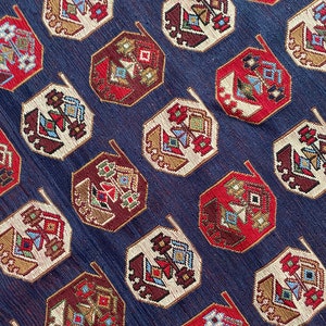 Op de afbeelding: Een close-up van een blauwe stof met een herhalend patroon van rode, witte en bruine geometrische vormen. De vormen zijn achthoekig en hebben een gestileerd ontwerp.