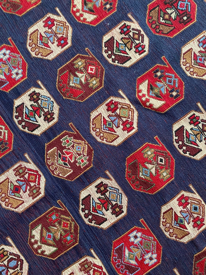 Op de afbeelding: Een close-up van een blauwe stof met een herhalend patroon van rode, witte en bruine geometrische vormen. De vormen zijn achthoekig en hebben een gestileerd ontwerp.