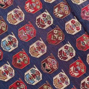 Op de afbeelding: Een close-up van een blauwe stof met een herhalend patroon van achthoekige vormen. De vormen zijn gevuld met een geometrisch ontwerp in tinten rood, wit, bruin en blauw.