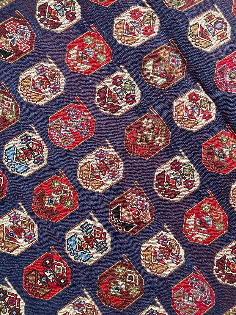 Op de afbeelding: Een close-up van een blauwe stof met een herhalend patroon van achthoekige vormen. De vormen zijn gevuld met een geometrisch ontwerp in tinten rood, wit, bruin en blauw.
