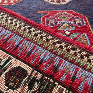 Op de afbeelding: Close-up van een rood, blauw en wit gepatroneerd tapijt met een franje rand. Het tapijt heeft een geometrisch ontwerp met ingewikkelde details.