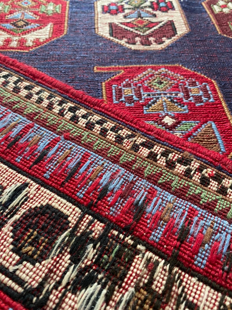 Op de afbeelding: Close-up van een rood, blauw en wit gepatroneerd tapijt met een franje rand. Het tapijt heeft een geometrisch ontwerp met ingewikkelde details.