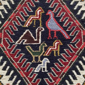 May include: A close-up of a colorful rug with a diamond-shaped pattern. The rug features a variety of stylized birds and horses in shades of red, blue, green, brown, and white. The background of the rug is a dark blue color.