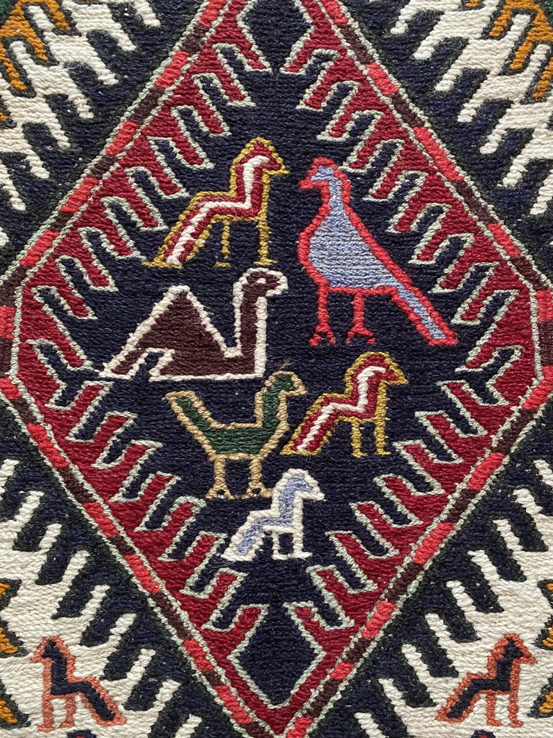 May include: A close-up of a colorful rug with a diamond-shaped pattern. The rug features a variety of stylized birds and horses in shades of red, blue, green, brown, and white. The background of the rug is a dark blue color.