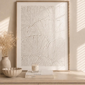 May include: A framed wall art piece featuring a textured, off-white relief of overlapping ginkgo leaves. The artwork is displayed in a thin, light-coloured wooden frame, showcasing a raised, sculptural surface with visible leaf veins and patterns.
