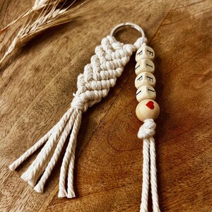 Peut inclure: Un porte-clés en macramé blanc avec un charme en perles de bois qui dit "FIFI" avec un cœur rouge sur la dernière perle.