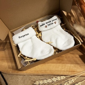 Peut inclure: Deux chaussettes de bébé blanches avec des revers, présentées dans une boîte en carton avec du papier déchiqueté. L'une dit "Surprise", l'autre "mini nous pour 2026". La boîte est posée sur une surface en toile de jute.