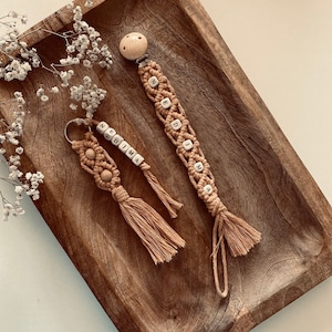 Könnte beinhalten: Handgefertigtes Set aus Babyaccessoires aus Makramee in Beige. Das Bild zeigt einen Schnullerclip und einen Schlüsselanhänger, beide mit Holzperlen, die Namen buchstabieren. Die Artikel werden auf einem Holztablett mit dekorativen Blumen präsentiert.
