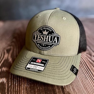 May include: Olive green and black trucker hat with the word "YESHUA" in a white and black patch. The hat has a curved brim and a mesh back. The patch also includes the words "EL SHADDAI" and "HU HAMELECH".