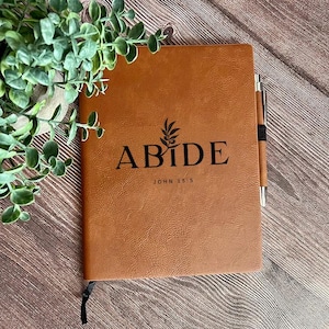 May include: A tan faux leather journal featuring the black text ABIDE with an olive branch graphic and the reference JOHN 15:5 printed on the front cover. A matching tan pen is secured in a side loop, and a black ribbon bookmark is visible.