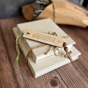 May include: A set of three white linen-covered books with a wooden bookmark and a wooden cross charm. The bookmark has the text "The Lord is my shepherd, I lack nothing He makes me lie down in green pastures, he leads me beside quiet waters he refreshes my soul Psalm 23".