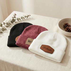 Puede incluir: Tres gorros de punto en negro, burdeos y blanco, cada uno con un parche de polipiel marrón. El gorro burdeos tiene las letras "BET" en el parche, y el blanco el nombre "Michelle".