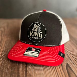 Könnte beinhalten: Eine schwarze, rote und weiße Baseballkappe mit dem Text "KING OF KINGS" auf der Vorderseite. Die Kappe hat einen roten Schirm, eine schwarze Krone und eine weiße Mesh-Rückseite. Das Richardson-Markenetikett ist sichtbar.