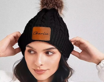 May include: Black cable knit beanie with a brown faux fur pom-pom. A brown leather patch on the front of the hat displays the name "Charlie". The hat is a winter accessory.