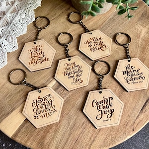Puede incluir: Una colección de llaveros hexagonales de madera con anillas de color bronce. Cada llavero presenta una cita inspiradora grabada en una fuente estilizada. Los llaveros están dispuestos sobre una tabla de madera.