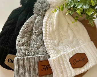 May include: Three knitted winter hats with pom poms and leather patches. The hats are gray, white, and black. The patches have the names "Tiffani", "Emily", and "CH" written on them.