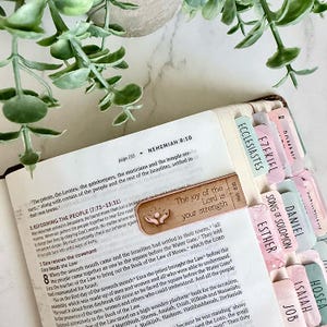 Może przedstawiać: Otwarta Biblia z drewnianą zakładką z wygrawerowanym tekstem The joy of the Lord is your strength oraz ikoną dłoni sięgających słońca. Strony Biblii zawierają kolorowe zakładki samoprzylepne z nazwami poszczególnych ksiąg.