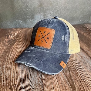 May include: Distressed navy blue baseball cap with a tan mesh back. The front features a brown leather patch with the word "PRAY" and a cross design. The cap has a pre-worn look with distressed details.