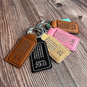Puede incluir: Una colección de llaveros de cuero en varios colores, incluyendo marrón, negro y rosa. Cada llavero presenta un versículo bíblico y un número de referencia. Los llaveros están unidos a llaveros plateados y se exhiben sobre una superficie de madera.