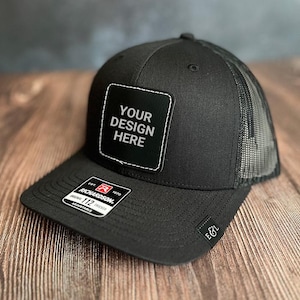 May include: Black trucker cap with a mesh back and a curved peak. The cap has a square patch with the text "YOUR DESIGN HERE". A small label on the front peak reads "RICHARDSON ORIGINAL 112 ADJUSTABLE".