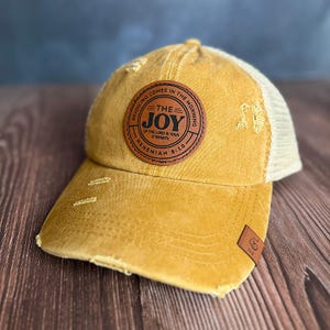 Könnte beinhalten: Eine senfgelbe, abgenutzte Baseballkappe mit Netzrücken. Ein runder brauner Lederaufnäher auf der Vorderseite zeigt den Text THE JOY OF THE LORD IS YOUR STRENGTH, REJOICING COMES IN THE MORNING und NEHEMIAH 8:10. Ein kleines E&L-Etikett befindet sich am Schirm.