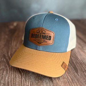 Puede incluir: Gorra de camionero azul, marrón y blanca con un parche de cuero. El parche dice "REDEEMED" con texto adicional. La visera es marrón, el panel frontal es azul y la parte trasera es de malla blanca. La gorra tiene una visera curva.