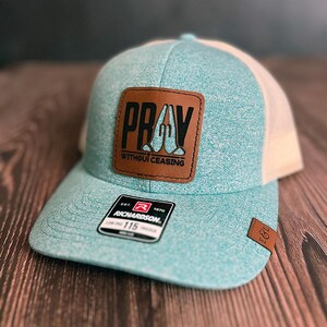 May include: A teal and tan trucker hat with a brown leather patch that reads "PRAY WITHOUT CEASING". The hat has a curved brim and a mesh back. A small tag with the brand name "Richardson" is visible.