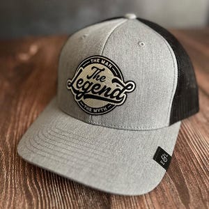 May include: Gray and black trucker hat with the text "The Man, The Legend, The Myth" in a circular patch. The hat has a gray front panel and brim, with a black mesh back. A small black tag with white text is on the side.