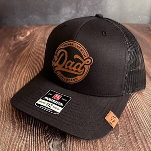 Könnte beinhalten: Schwarze Trucker-Kappe mit Mesh-Rückseite und gebogenem Schirm. Die Kappe hat einen braunen Lederaufnäher mit den Worten "Dad" und "The Man, The Myth, The Legend" in einem kreisförmigen Design. Ein kleines Etikett mit "E&L" befindet sich an der Seite.