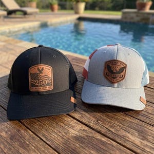 May include: Two baseball caps, one black and one light grey, each featuring a brown leather patch. The black cap displays an eagle, a flag, and the text 250 YEARS ONE NATION UNDER GOD. The grey cap features an eagle and the text ONE NATION UNDER GOD 250 YEARS 1776 2026.