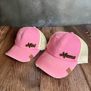 Puede incluir: Dos gorras de béisbol rosas con detalles desgastados. Una gorra dice "Mini" y la otra "Mama" en letras marrones. Ambas gorras tienen una parte trasera de malla beige y una pequeña etiqueta de cuero marrón.