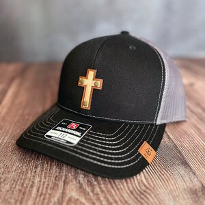 May include: Black and gray trucker hat with a light brown leather cross patch. The hat features a curved brim, white stitching, and a mesh back. The Richardson brand label is visible.