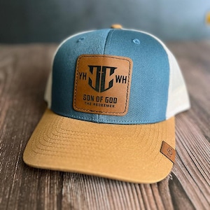 Könnte beinhalten: Blau-beige Baseballkappe mit weißem Mesh-Rücken. Die Kappe hat einen braunen Lederaufnäher mit dem Text "SON OF GOD THE REDEEMER". Der Schirm ist beige und die Krone blau.