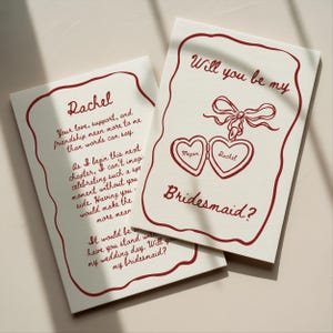 May include: Two cream-colored cards with red text and borders. One card reads "Rachel" with a personal message. The other says "Will you be my Bridesmaid?" with two heart-shaped lockets.