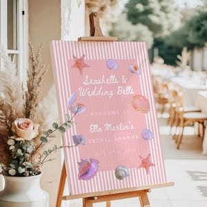 May include: A pink and white striped welcome sign on an easel. The sign reads "Seashells & Wedding Bells" and welcomes guests to Ella Martin's bridal shower on July 27th, 2026. Seashell and starfish illustrations decorate the sign.