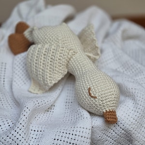 Può includere: Un giocattolo a forma di oca all'uncinetto fatto a mano, bianco sporco, con dettagli marroni. L'oca presenta un lungo collo, ali e un becco e zampe dettagliati. Il giocattolo è appoggiato su una coperta bianca strutturata.
