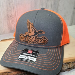 May include: A gray and orange trucker hat with a brown leather patch that says "Only Ducks". The patch features a flying duck. The hat is adjustable and has a Richardson 112 label.