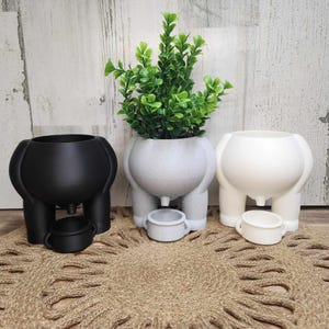 May include: Three 3D printed planters shaped like cows, one black, one gray, and one white. Each planter has a small pot underneath for watering. The gray planter has a green plant inside.