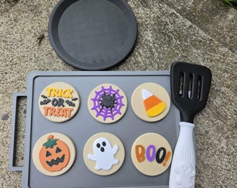 May include: A gray rectangular griddle with six round Halloween-themed inserts featuring a pumpkin, ghost, spider web, candy corn, and text reading Trick or Treat and Boo. A black and white spatula with a ghost design sits next to the griddle.