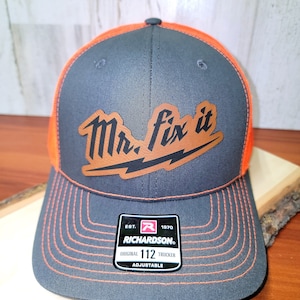 May include: A gray and orange trucker hat with an adjustable strap. The hat features a brown leather patch with the text "Mr. Fix It" in a stylized font with a lightning bolt.