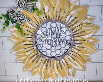 May include: A yellow sunflower wreath with a distressed white paint finish. The wreath has a wire mesh center with the words "Hello Sunshine" written in white. The wreath is decorated with a black and white bow and green leaves.