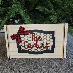 Puede incluir: Un letrero de madera blanco con un marco negro y un patrón de panal. El letrero dice "The Carlins" en letras rojas con un lazo rojo y negro a cuadros.