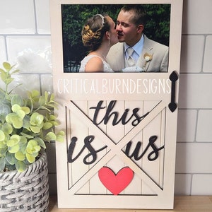 May include: A white wooden sign with a picture frame in the top half. The bottom half has a white wooden door with a red heart and the words "this is us" in black.