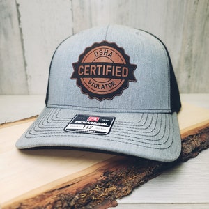 May include: A gray and black trucker hat with a brown leather patch that says "OSHA Certified Violator".