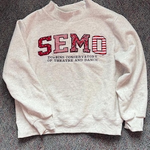 Puede incluir: Una sudadera gris claro con cuello alto. La palabra "SEMO" está en el pecho en letras rojas con diseños estampados. Debajo, el texto "DOBBINS CONSERVATORY OF THEATRE AND DANCE" es en rojo.