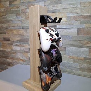 Könnte beinhalten: Ein Holzständer für Gamecontroller mit drei Controllern. Der Ständer ist aus hellem Holz gefertigt und hat eine schwarze Halterung. Die Controller sind weiß mit roten Spritzern und schwarz mit bunten Designs.