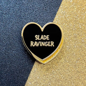 Puede incluir: Un pin de esmalte en forma de corazón con un centro negro y un borde dorado. El pin presenta las palabras "SLADE RAVINGER" en letras blancas. El fondo es una división de azul marino y purpurina dorada.