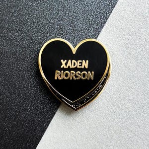 May include: A black heart-shaped pin with gold trim and the name "XADEN RIORSON" in gold lettering. The pin is set against a black and white background.