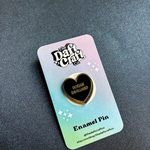 May include: A black heart-shaped enamel pin with gold trim and the name "Dorian Havilliard" in white text. The pin is on a colorful card with the text "Enamel Pin" and the shop's logo and social media handles.