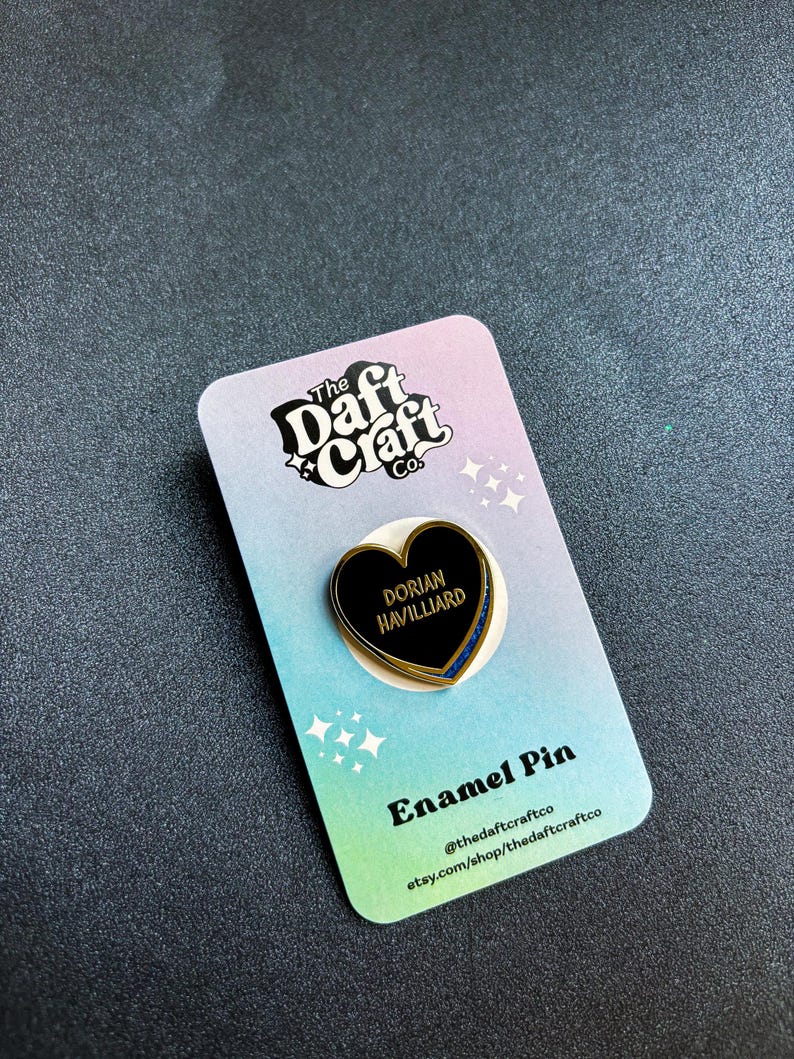 May include: A black heart-shaped enamel pin with gold trim and the name "Dorian Havilliard" in white text. The pin is on a colorful card with the text "Enamel Pin" and the shop's logo and social media handles.